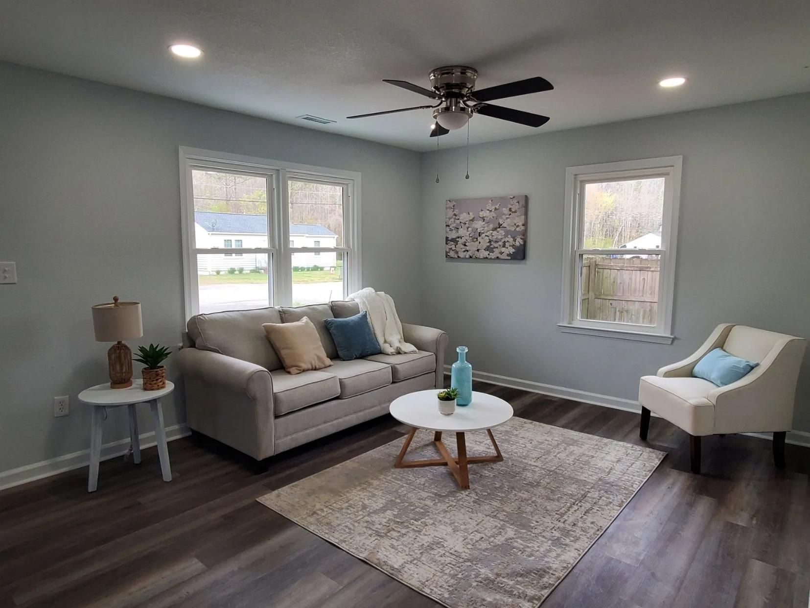 Bright living room with gray walls, beige sofa, white chair, round coffee table, and two windows.