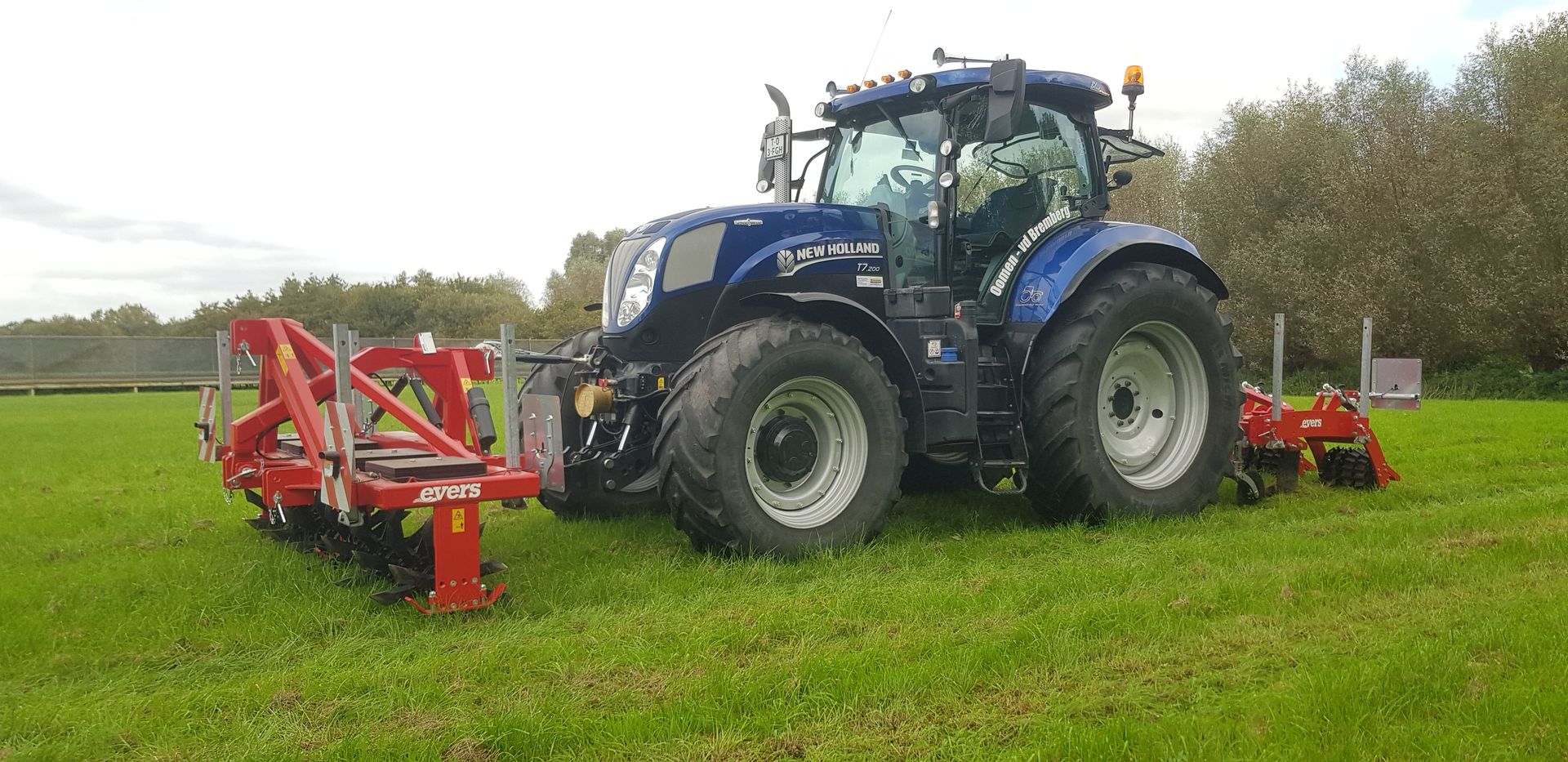Een blauwe tractor maait gras met rode landbouwmachines op een groen veld.