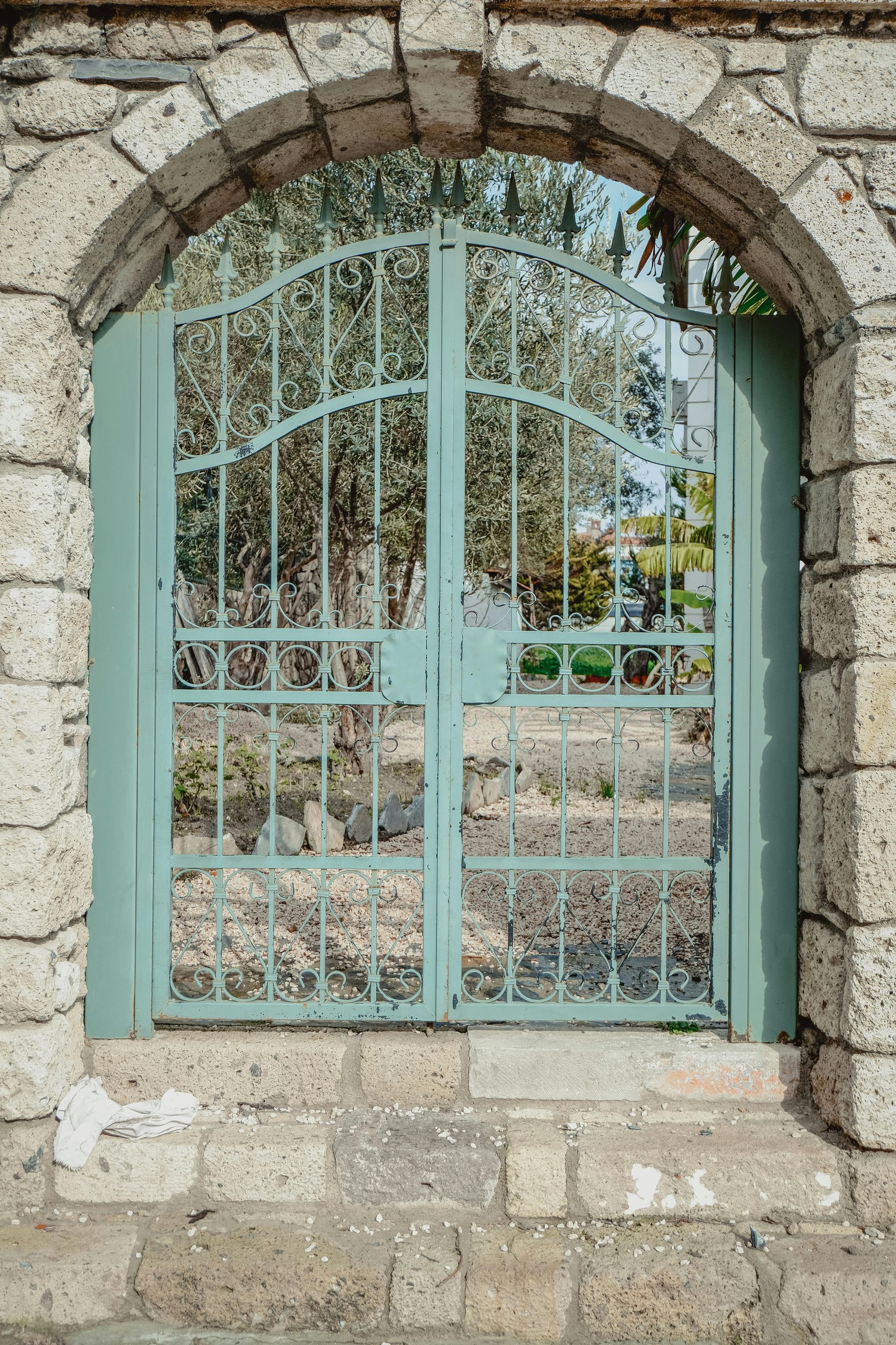 Blue-green metal gate set in a stone archway. Background of green trees visible through the gate.