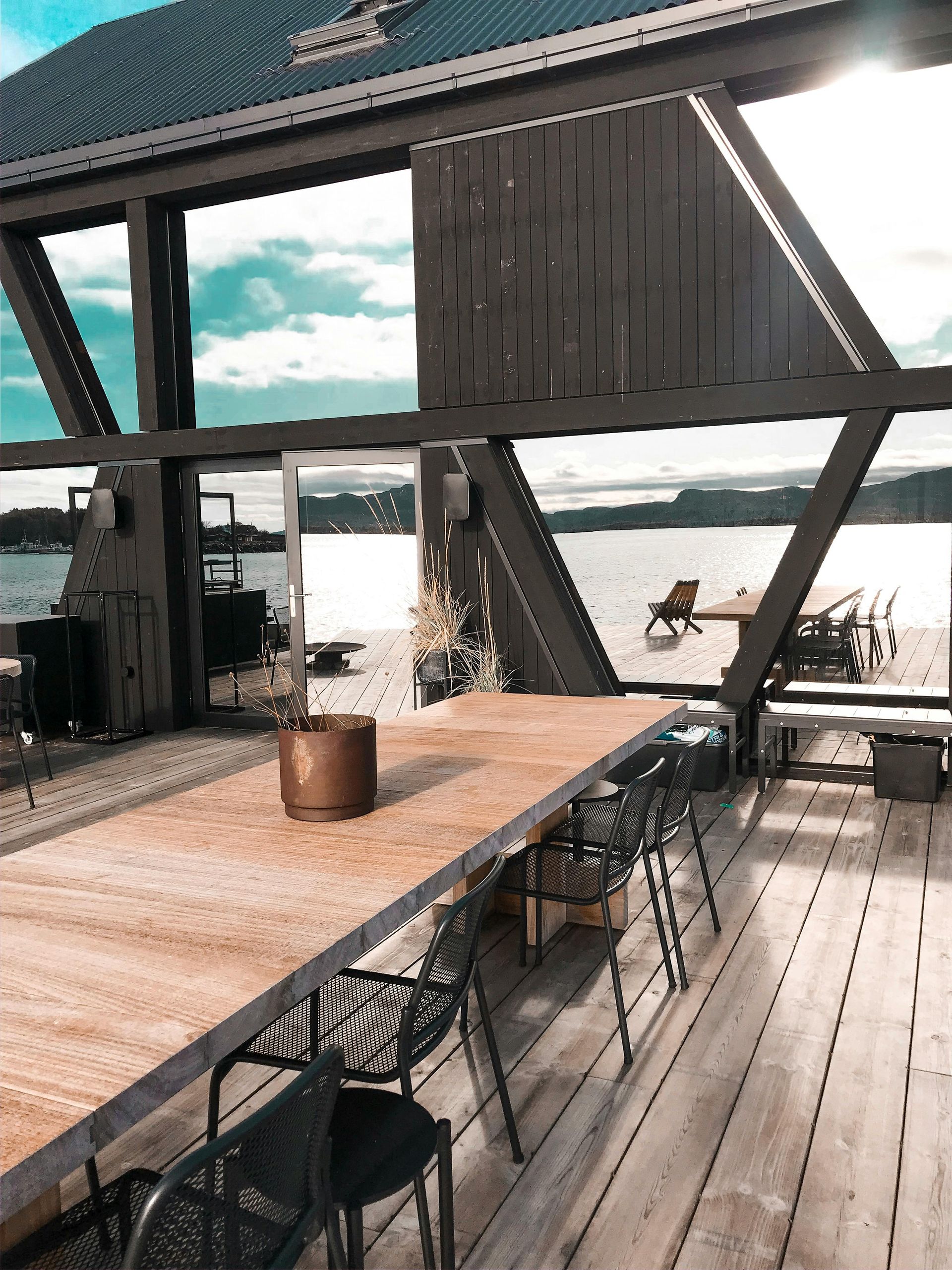 Outdoor dining area with a long wooden table, black chairs, and a lake view.