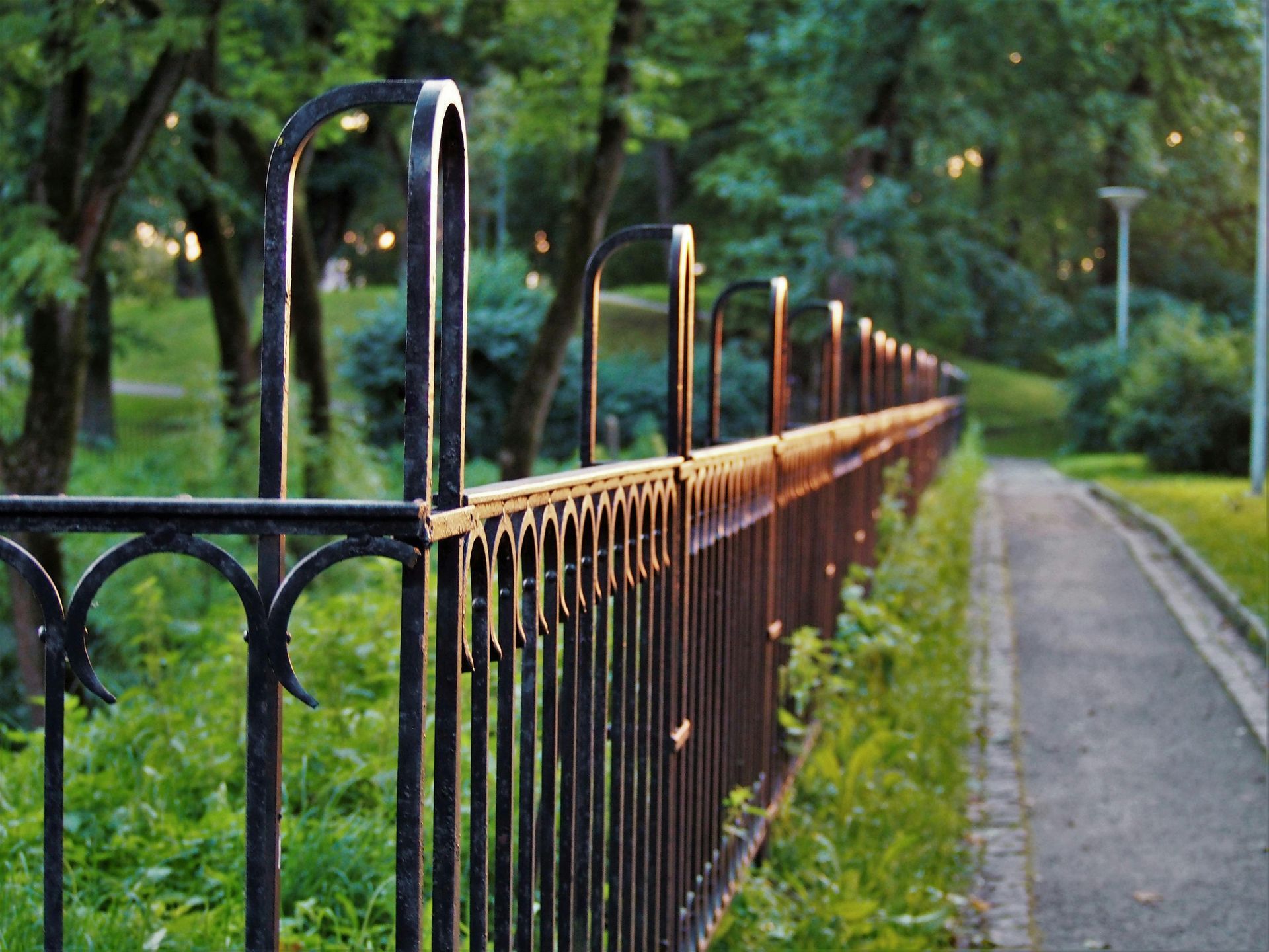 Black metal fence along a paved path in a park with trees and grass, bathed in sunlight.