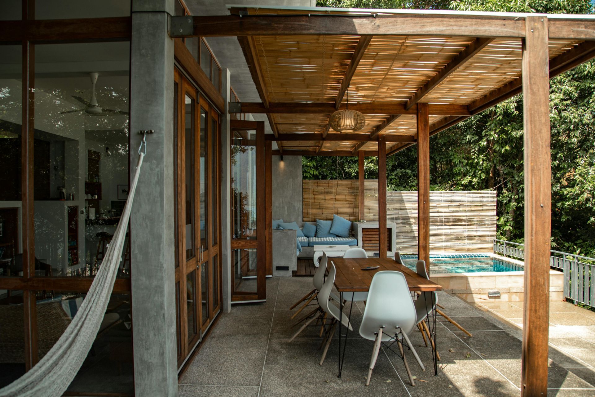 Patio with wooden pergola, dining table, pool, and hammock next to a house with large windows.