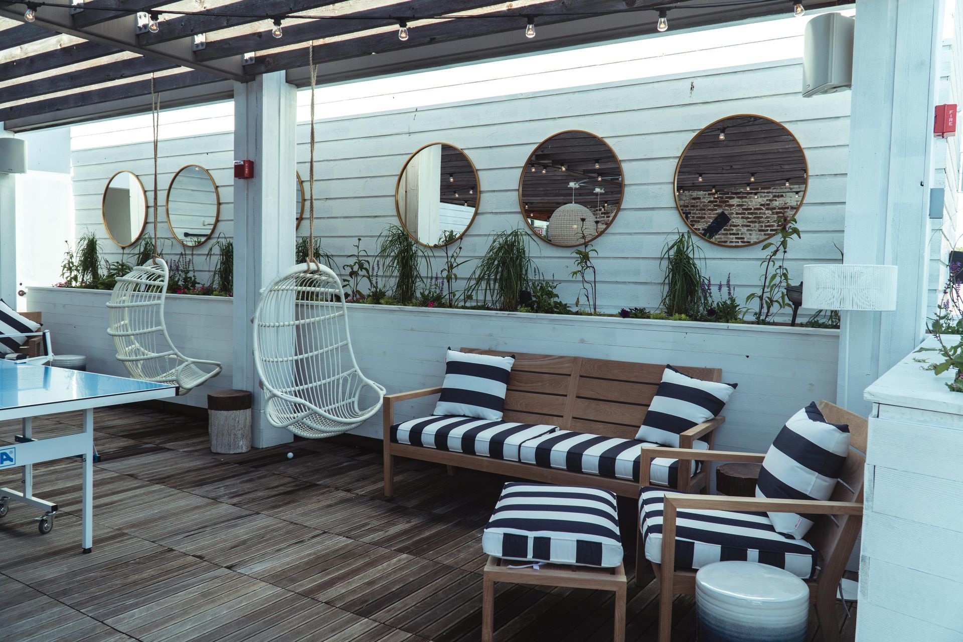 Outdoor seating area with striped cushions, hanging chairs, and round mirrors on a white wall.
