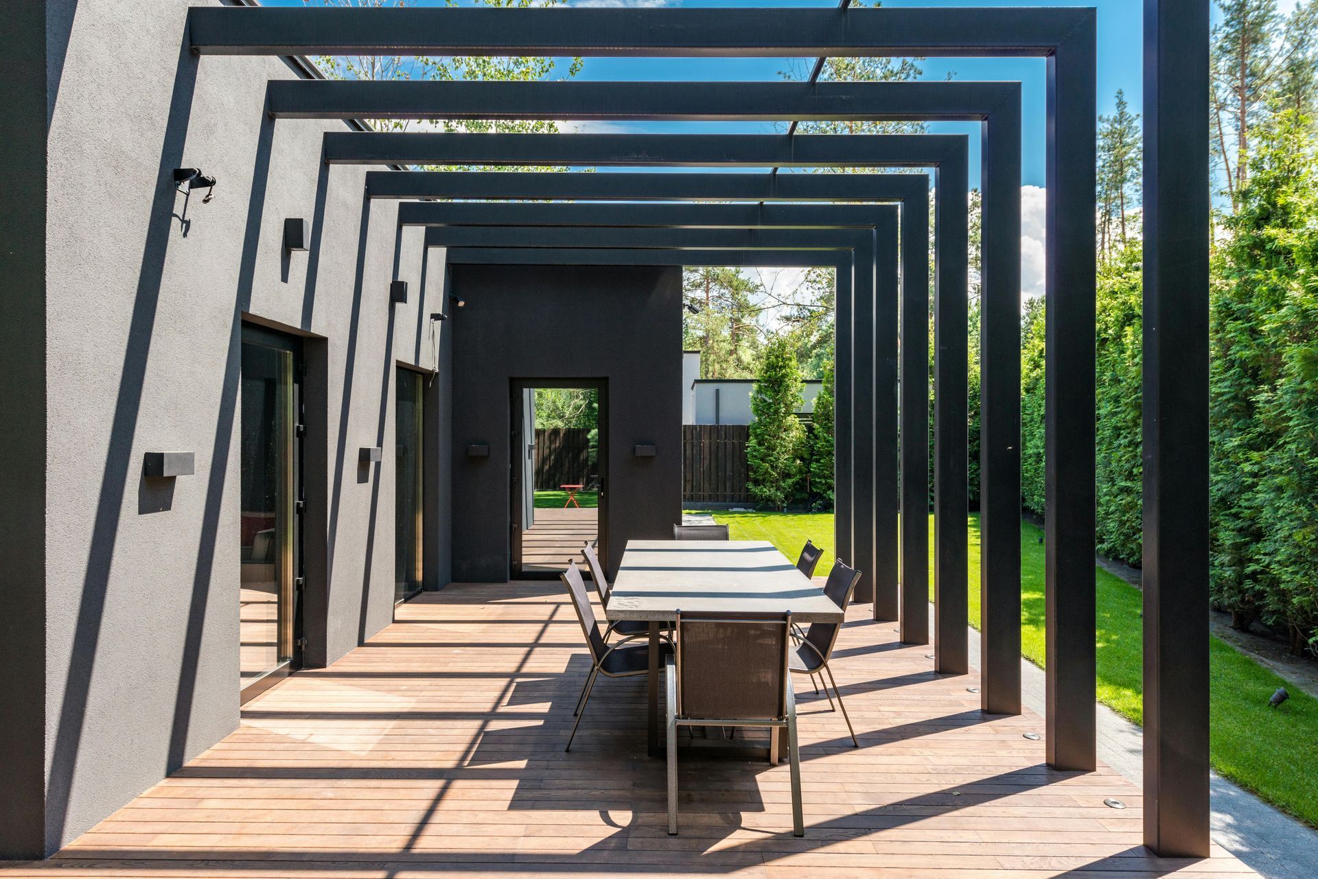 Black pergola over outdoor dining table on a wooden deck. Sunlight casts shadows on the wall.