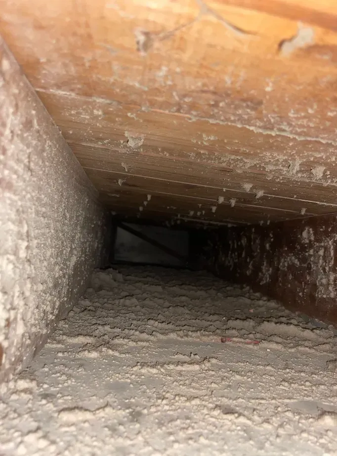 Dusty crawlspace duct interior with white debris along the floor and wood framing overhead