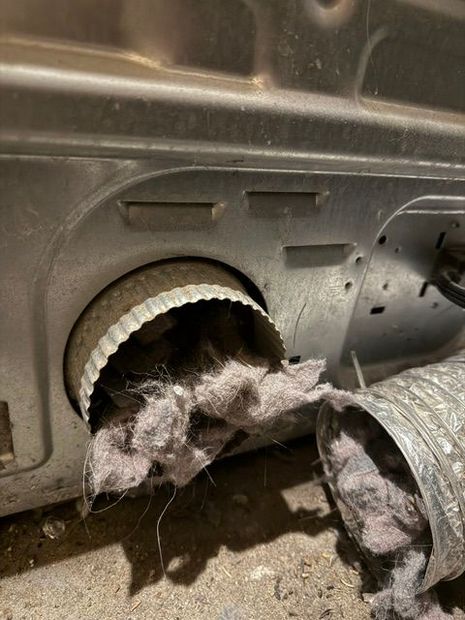 Cat hair clogged in a dryer vent opening, with lint piled on the floor and a detached hose nearby