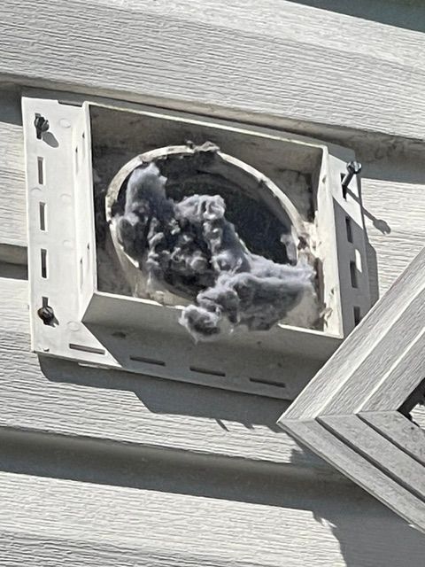 Damaged exterior vent with torn gray insulation or debris, mounted on light gray siding.