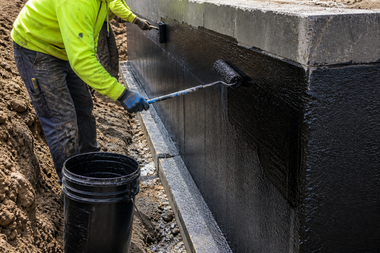 Worker spraying black coating on a concrete wall beside a bucket outdoors