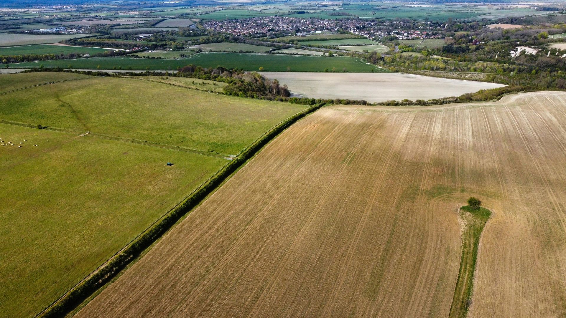 Aerial view of green and tan farmland with a pale pond and distant village under a clear sky