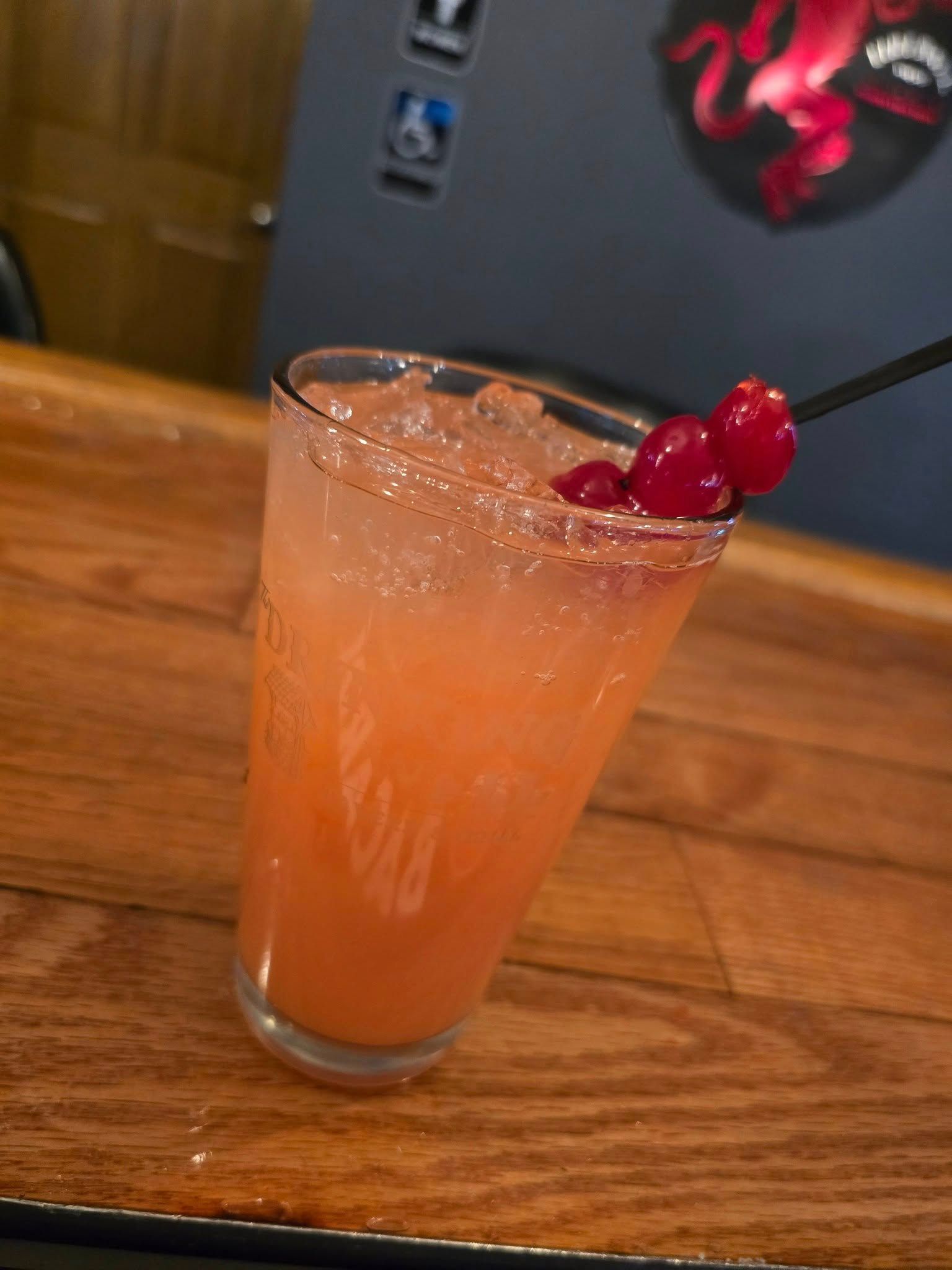 A glass of orange liquid with a cherry on top is on a wooden table.