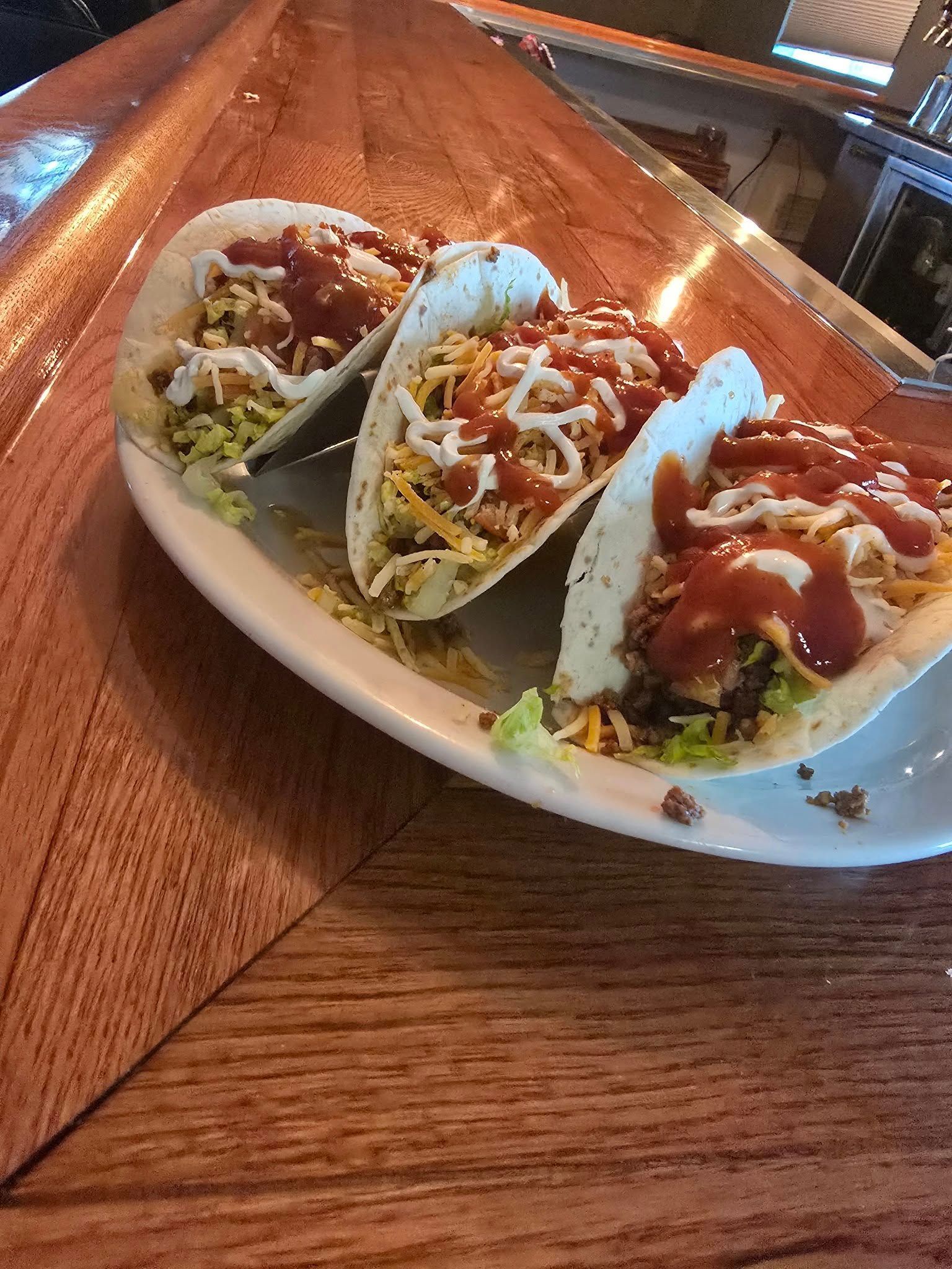 Three tacos are sitting on a white plate on a wooden table.