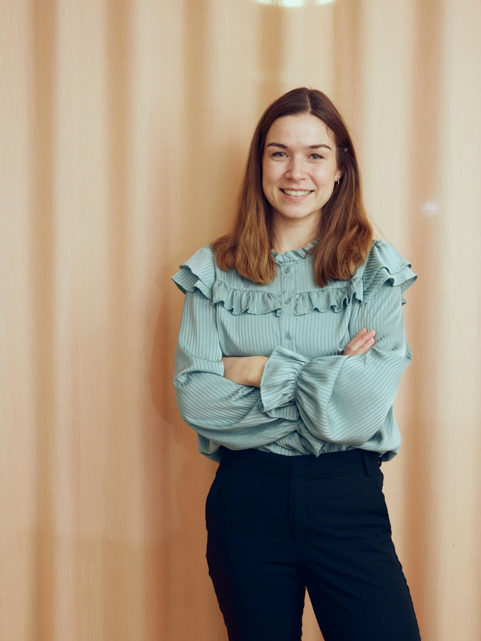 Eine Frau in einer grünen Bluse und schwarzen Hosen steht mit verschränkten Armen da.