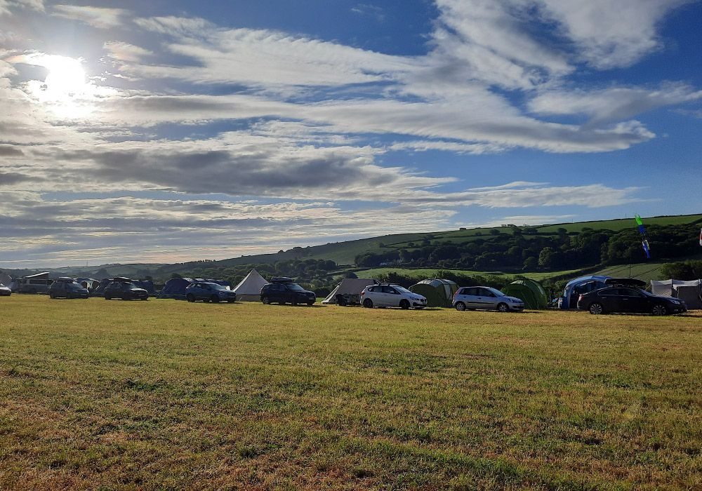 Cars and tents in a grassy field under a cloudy blue sky, hills in the background.