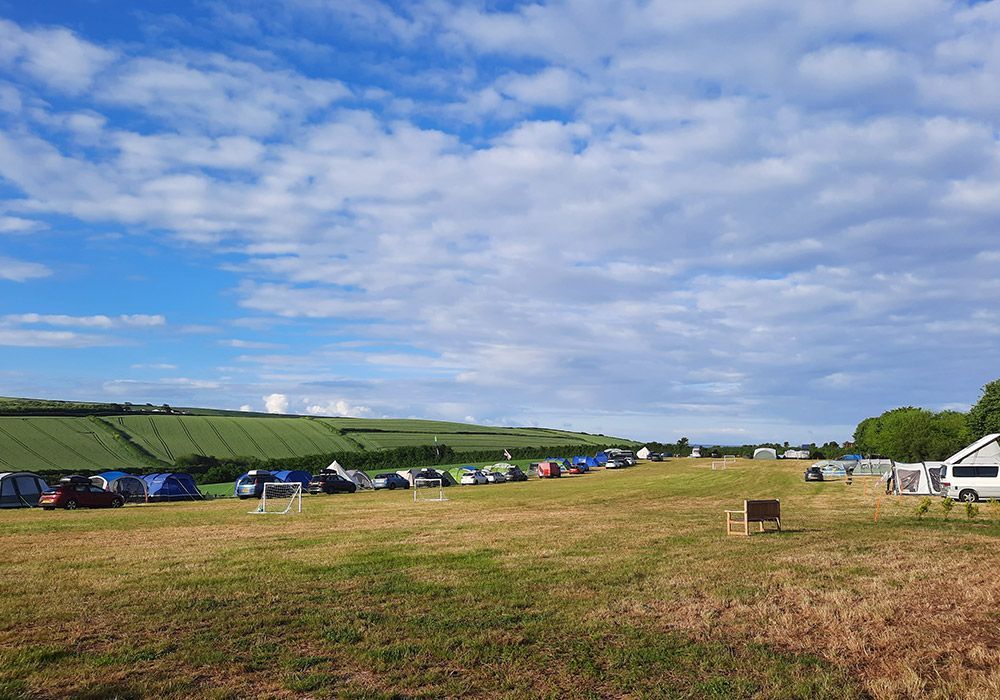 Camping field under a blue sky, with numerous tents and green rolling hills in the background.