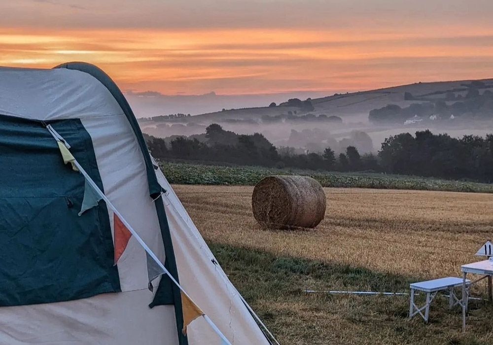 Camping tent in a field with a hay bale at sunrise. Orange and pink sky, misty hills in background.