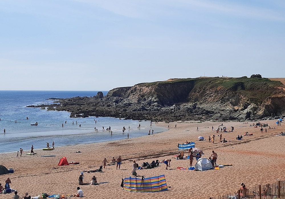 Beach with people enjoying a sunny day; sand, ocean, rocky cliffs, blue sky.