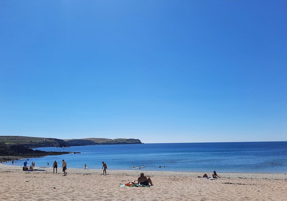 Beach scene with people relaxing, swimming, and enjoying a sunny day; blue sky and water.