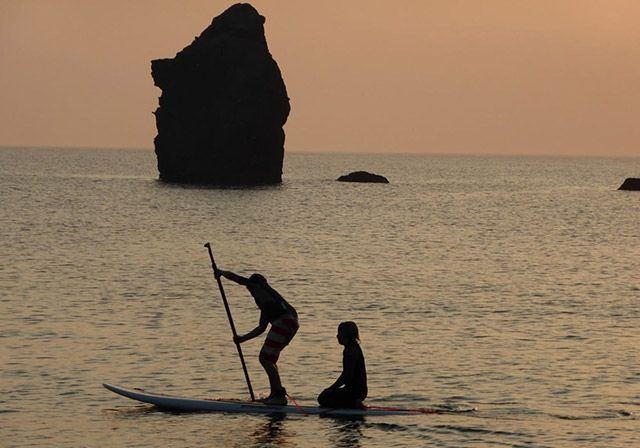 Silhouette of two people paddleboarding at sunset, with a large rock in the ocean.