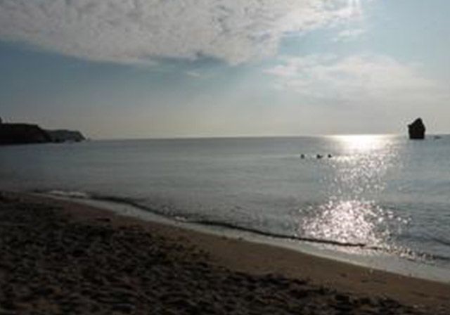 Beach with calm water reflecting sunlight; cliffs on either side; a rock formation in the distance.