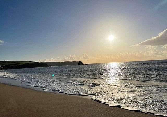Sunlit beach scene with waves, shoreline, and a distant rock formation.