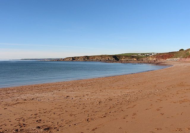 Sandy beach with red-brown sand, calm blue water, and a rocky coastline under a clear blue sky.