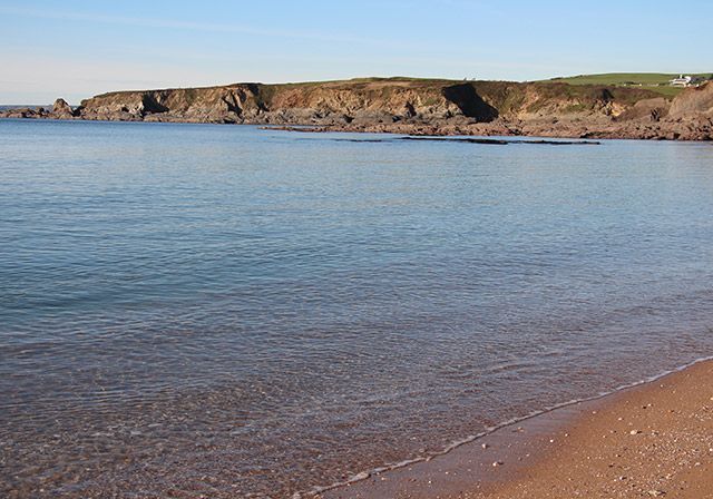 Calm ocean water meets a sandy beach. Rocky cliff in the background under a blue sky.