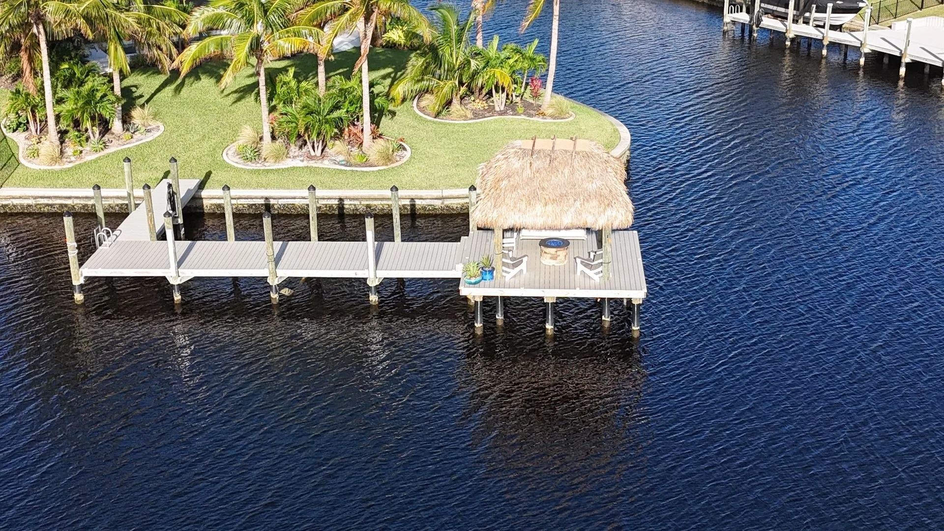 Waterfront dock beside a palm-lined tropical lawn with thatched-roof huts and dark blue water