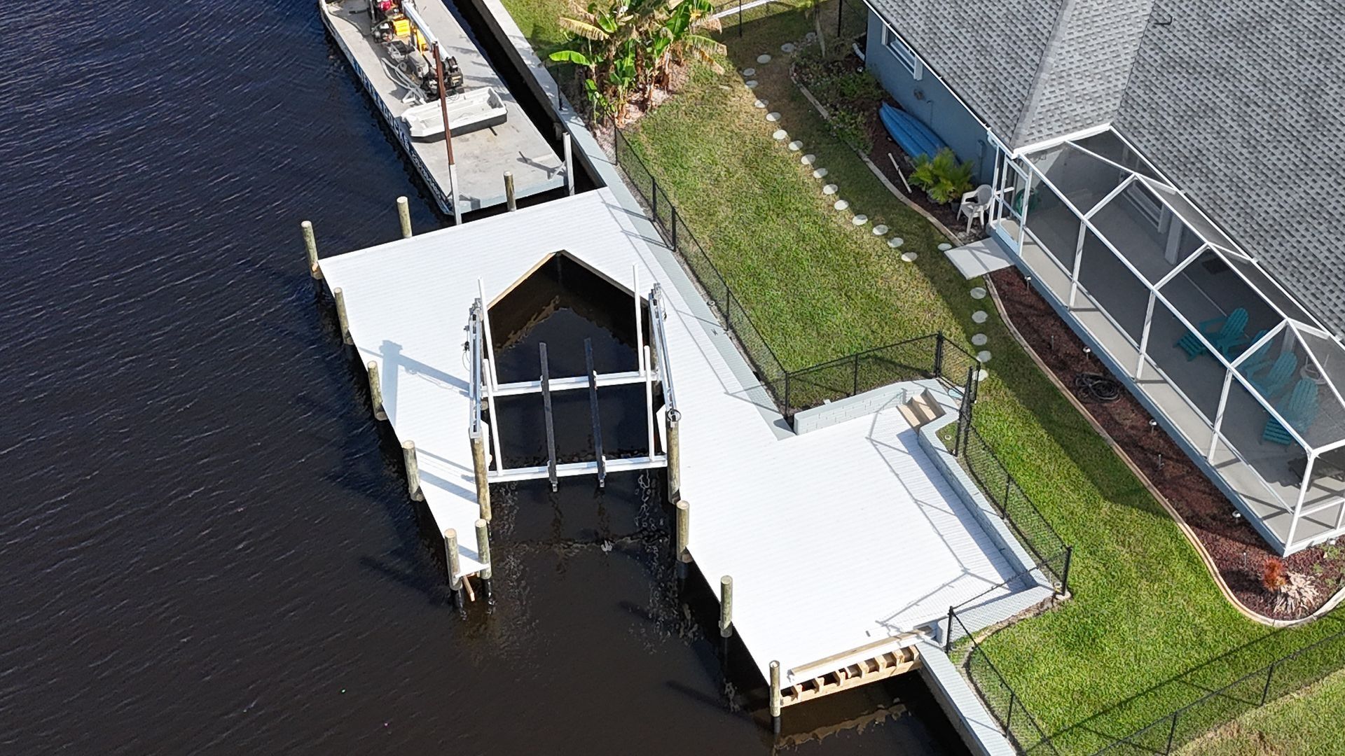 Aerial view of a white dock extending into dark water beside a greenhouse and grassy shoreline.