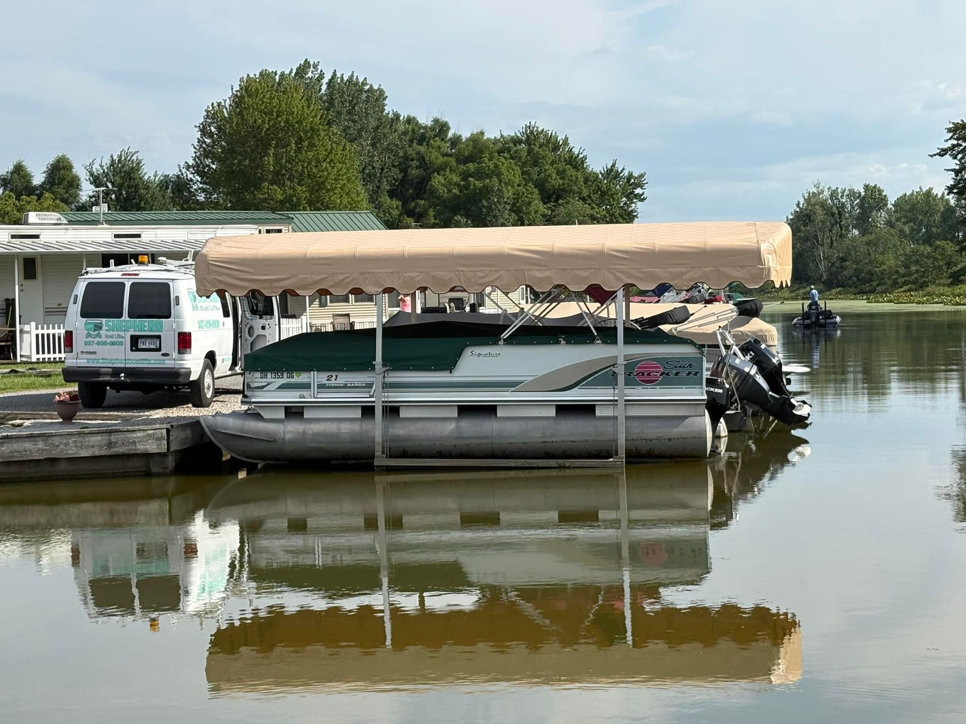 Pontoon boat with tan canopy tied at flooded dock, with white van on the left and trees reflected in calm water.