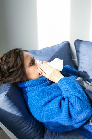 Person lying on a blue couch, holding a tissue to their face and wearing a blue sweater.