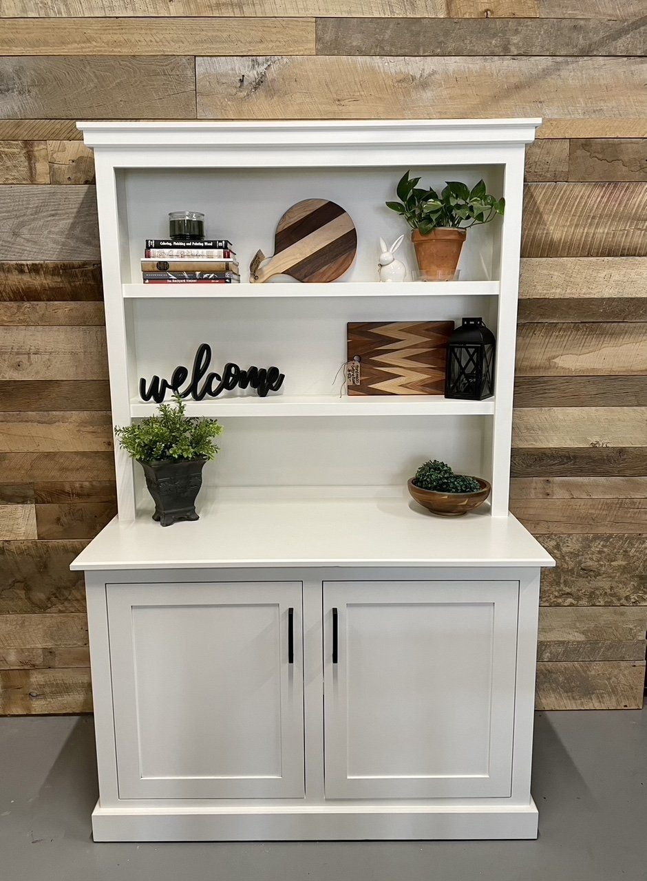 A white bookshelf with a welcome sign on it is sitting in front of a wooden wall.