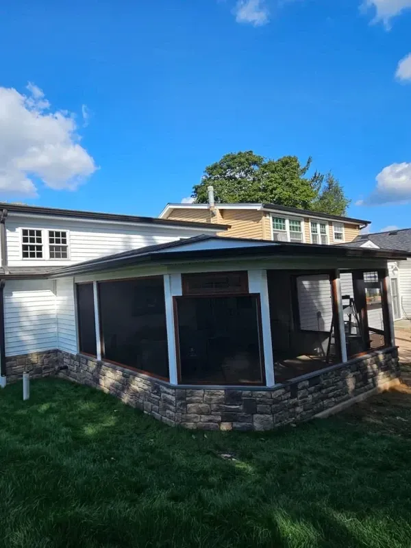 Screened-in porch with stone facade and white trim, attached to a house with a blue sky background.
