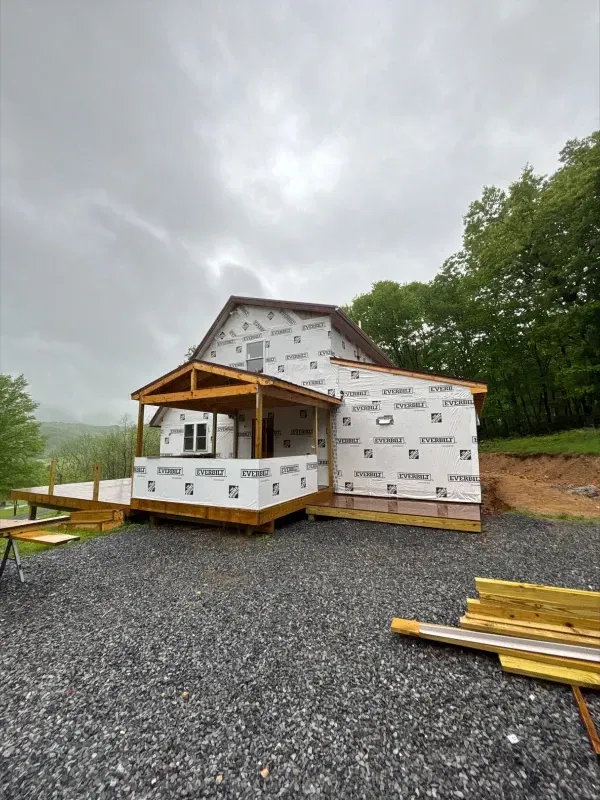 House under construction with a deck, porch, and vapor barrier wrap. Gray gravel ground and overcast sky.