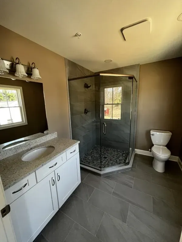 Bathroom with a white vanity, a corner glass shower, and a toilet. Gray tile floor and brown walls.