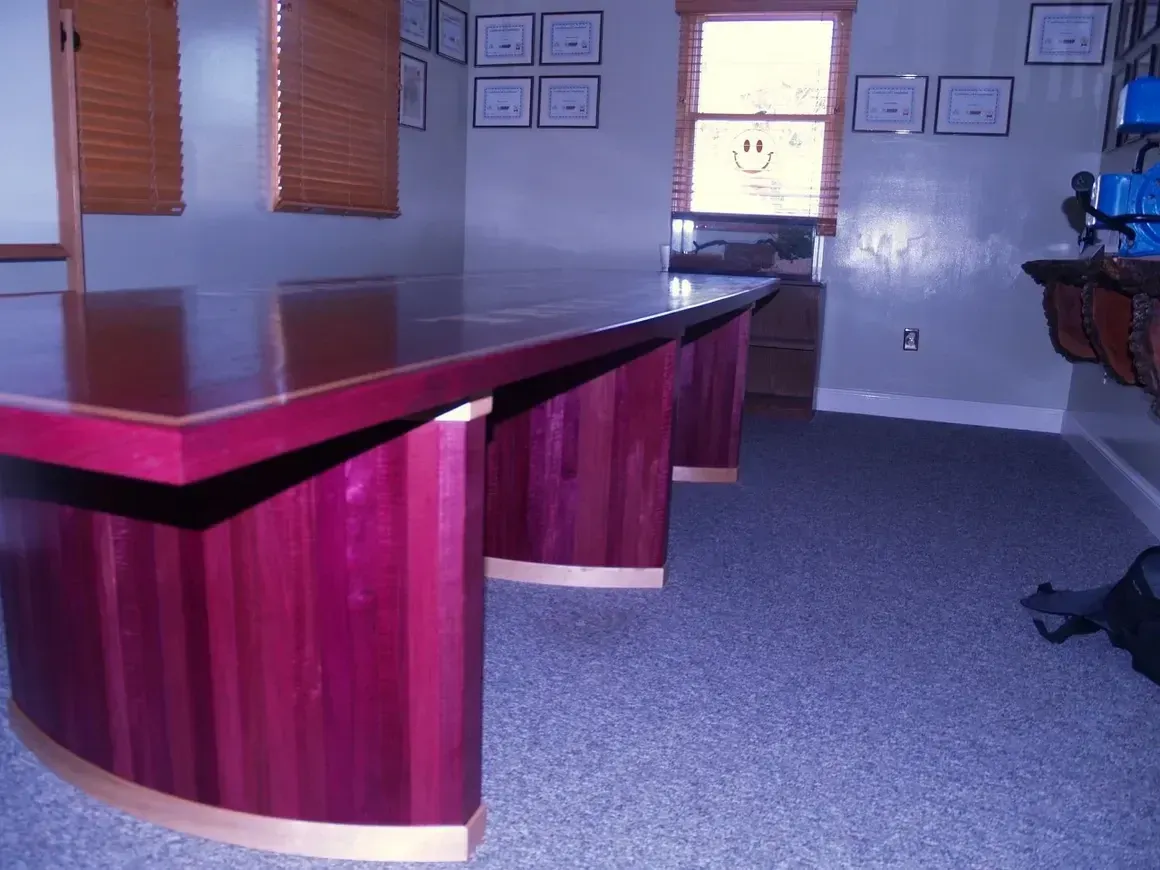 Conference room with large, purple wooden table.  Brown blinds, window, blue carpet, awards on wall.