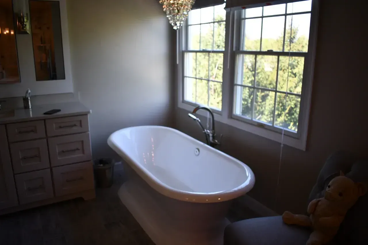 Bathroom with white pedestal tub, window, and vanity. Neutral walls, chandelier, and a teddy bear.