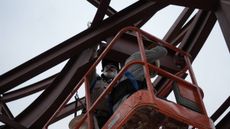 Worker in a red aerial lift inspecting dark steel bridge beams overhead
