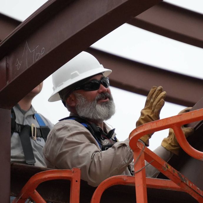 Construction worker in white hard hat and sunglasses, directing steel beam placement from an orange lift basket.