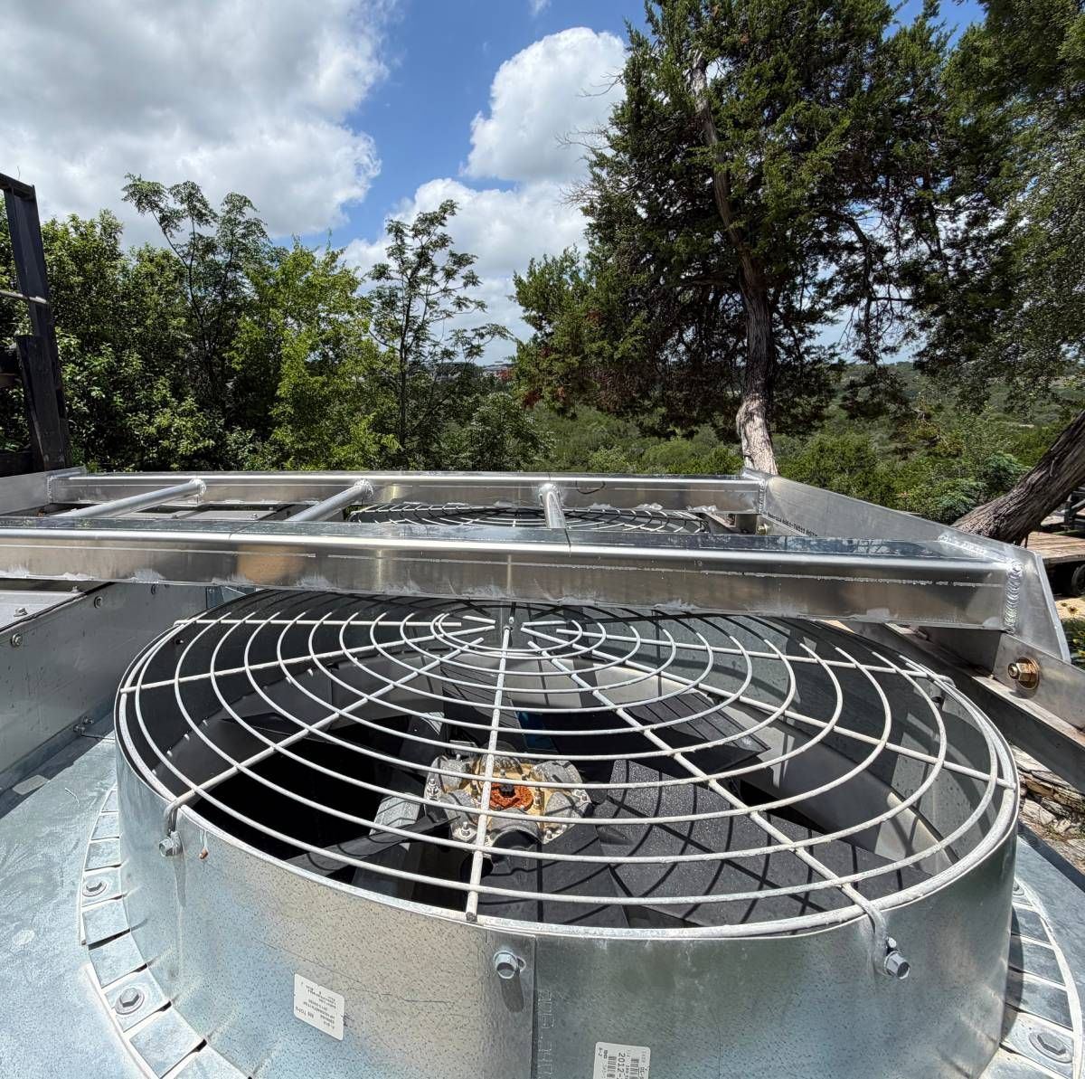 Outdoor HVAC fan unit on a rooftop with trees and cloudy sky in the background