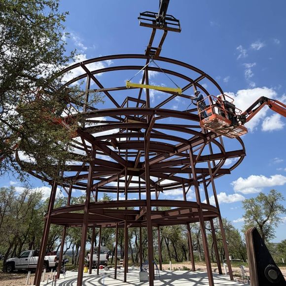 Steel-framed circular structure under construction, with a worker in a boom lift against a blue sky.