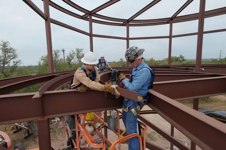 Two workers on a steel beam wearing harnesses, bolting a roof frame under construction