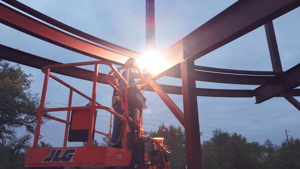 Worker on a red lift assembling a steel frame at sunset, with the sun shining through the beams