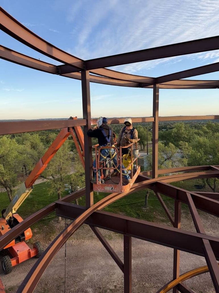 Workers on a steel tower platform at sunset, surrounded by forest and open sky.