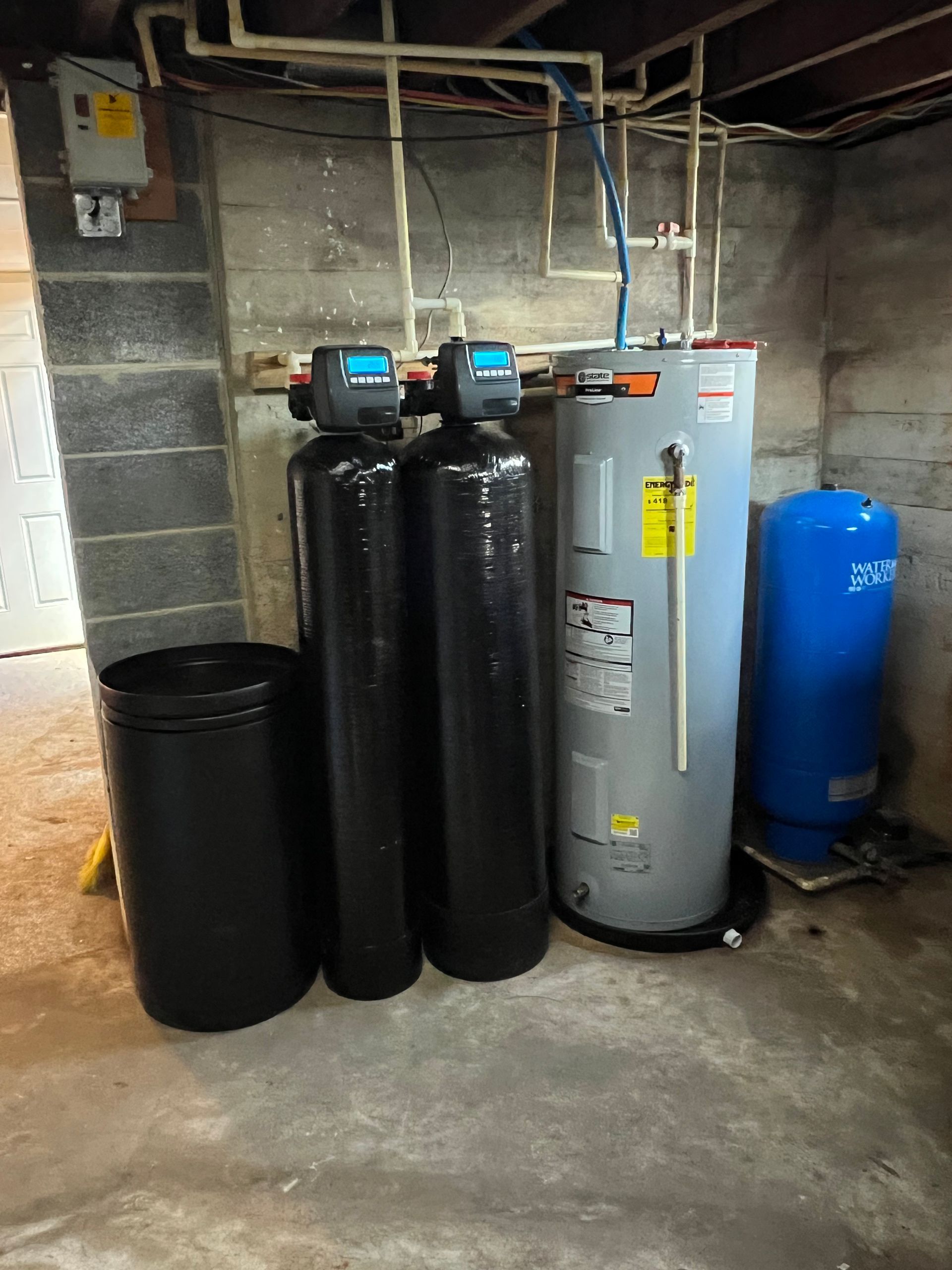 Water softener and filtration tanks in a basement utility area, with pipes and a blue pressure tank.