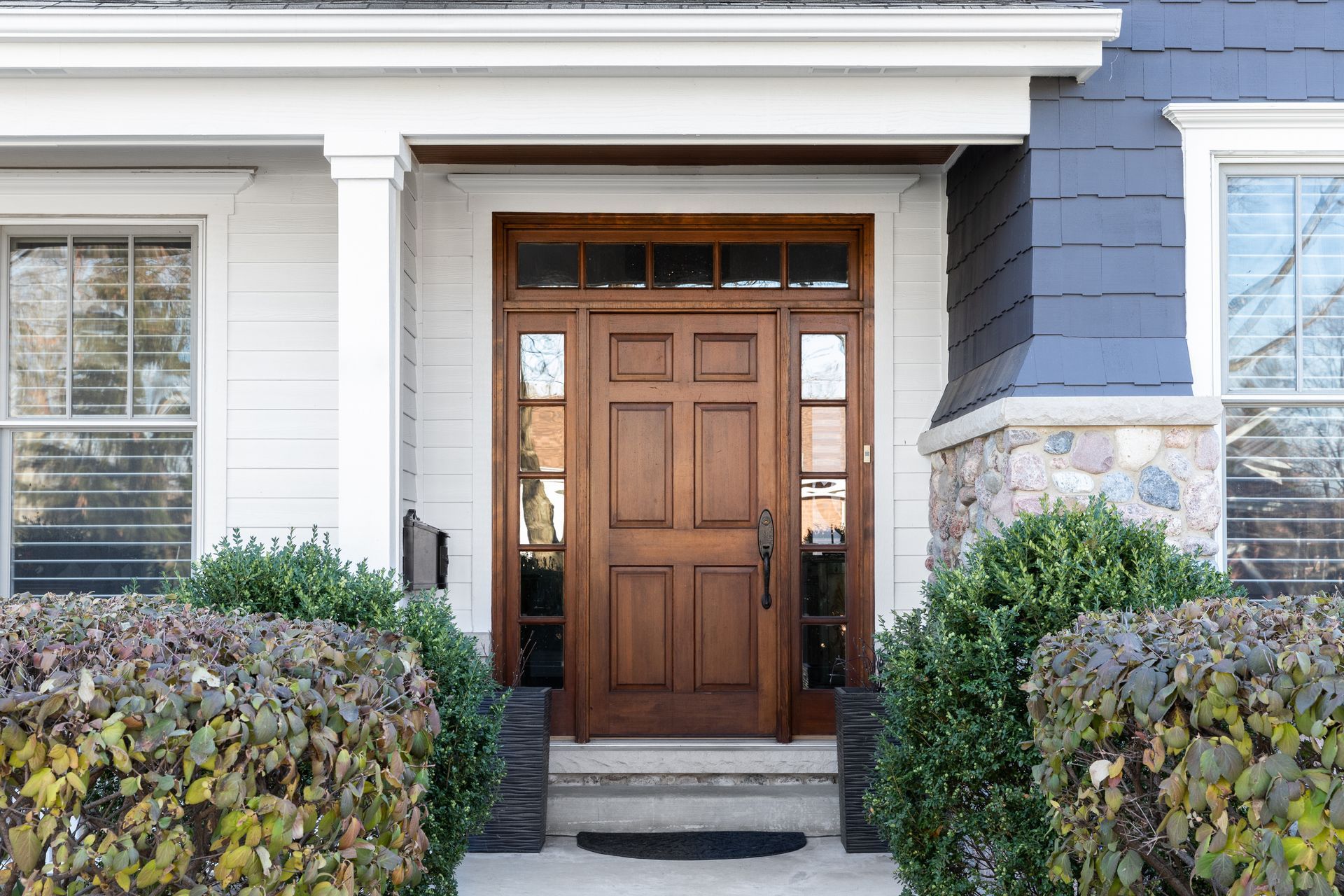 Wooden front entry door with glass sidelights, framed by white siding, stone accents, and shrubs. Wooden front entry door with glass sidelights, framed by white siding, stone accents, and shrubs.