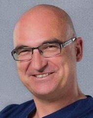 A bald man wearing glasses and a blue scrub is smiling.