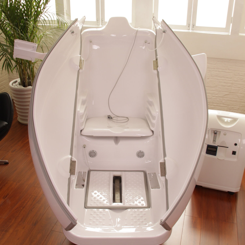 White oval spa pod with a seat and foot basin in a bright room, next to a small plant and white device.