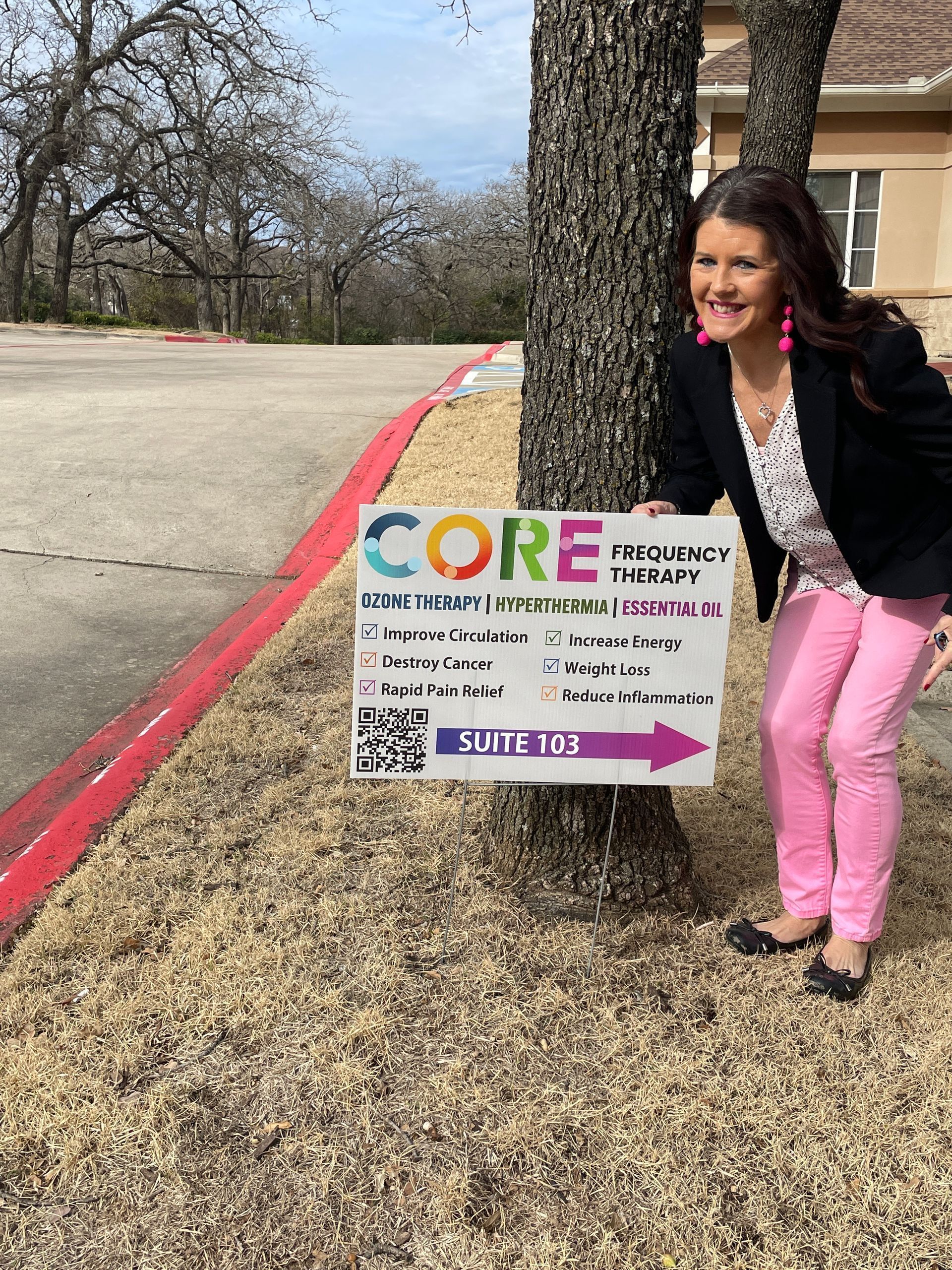 Woman kneeling beside a CORE campaign sign outdoors on a sidewalk by a building and parking lot
