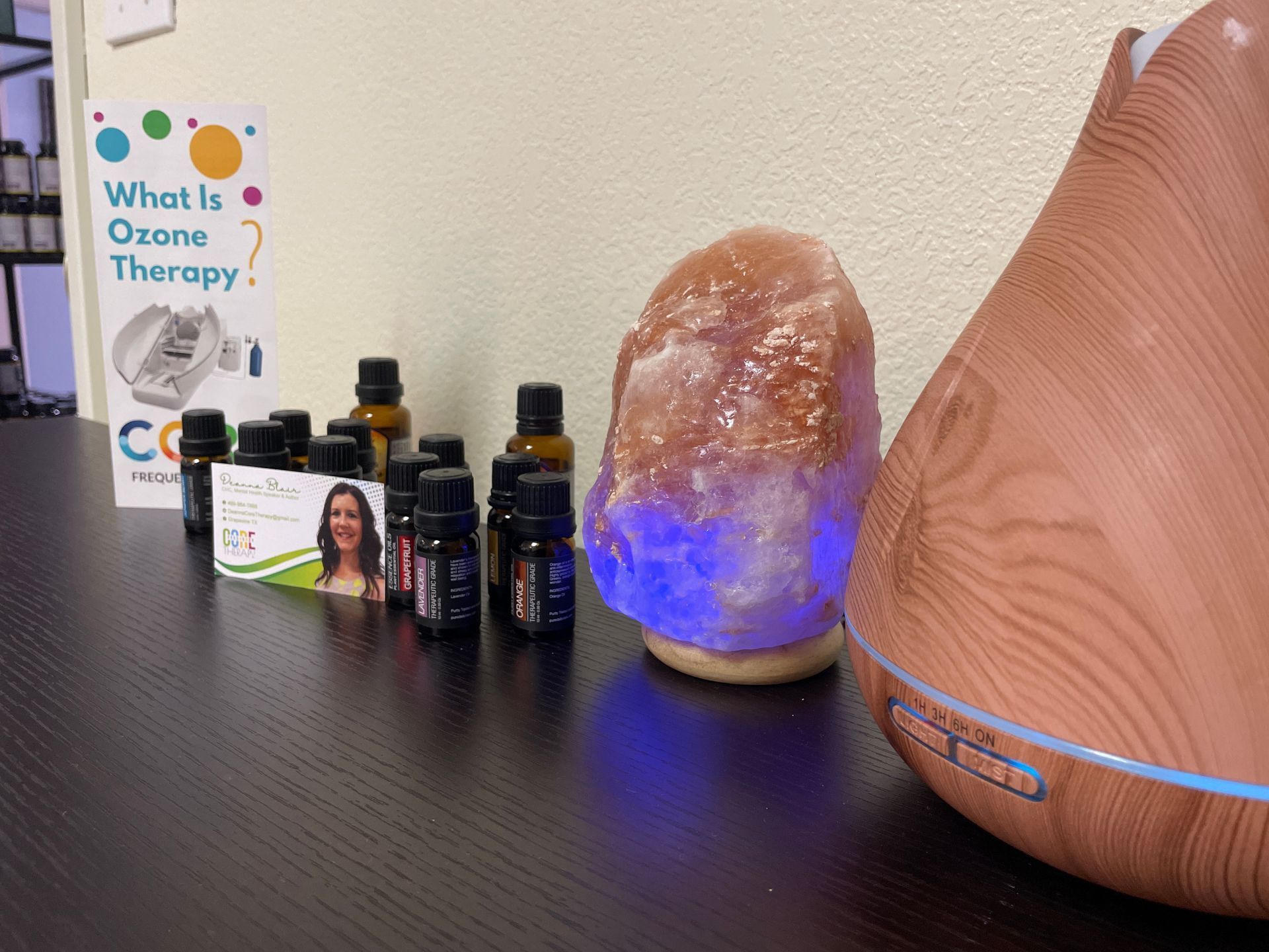 A tabletop display with essential oil bottles, a blue-lit salt lamp, and a wood diffuser beside a “What’s Ozone Therapy?” card