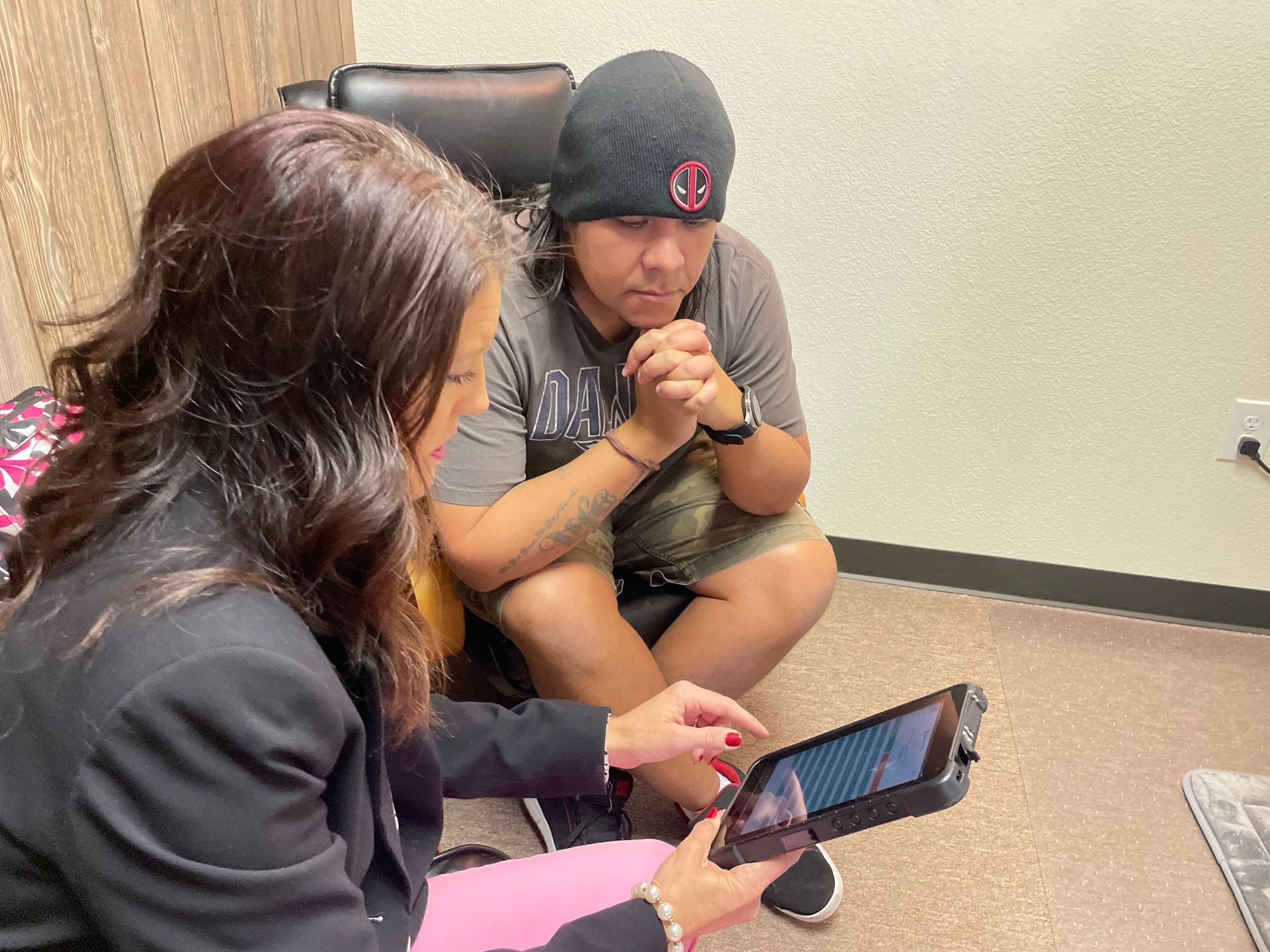 Two people sit on a floor discussing a tablet in a small office.